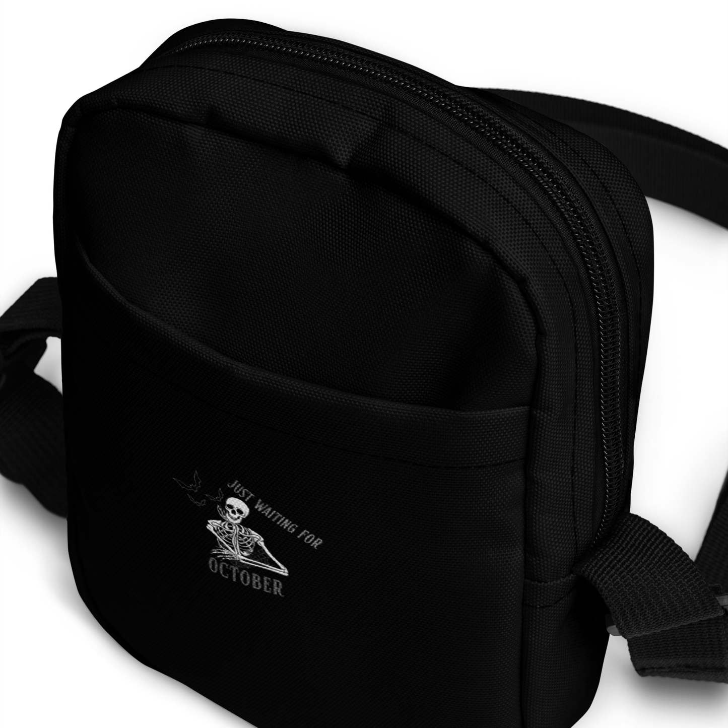 This is a black crossbody bag that has a bored skeleton with his head resting in his hand and he has spider webs and bats around him and the text reads just waiting for October