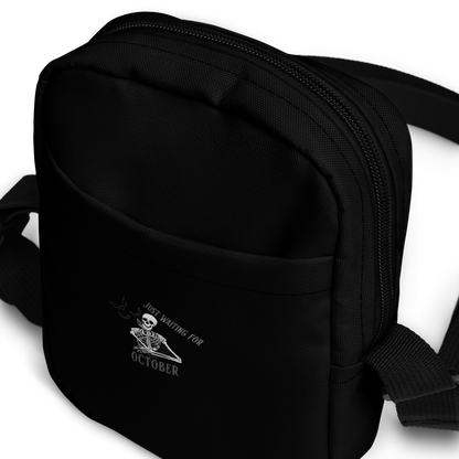 This is a black crossbody bag that has a bored skeleton with his head resting in his hand and he has spider webs and bats around him and the text reads just waiting for October