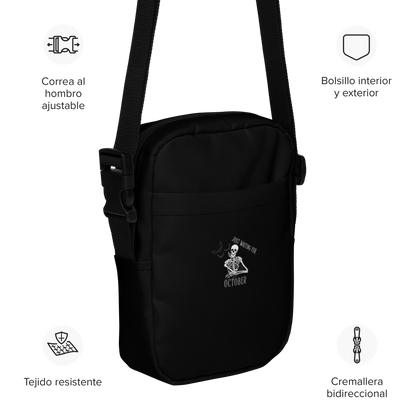 This is a black crossbody bag that has a bored skeleton with his head resting in his hand and he has spider webs and bats around him and the text reads just waiting for October