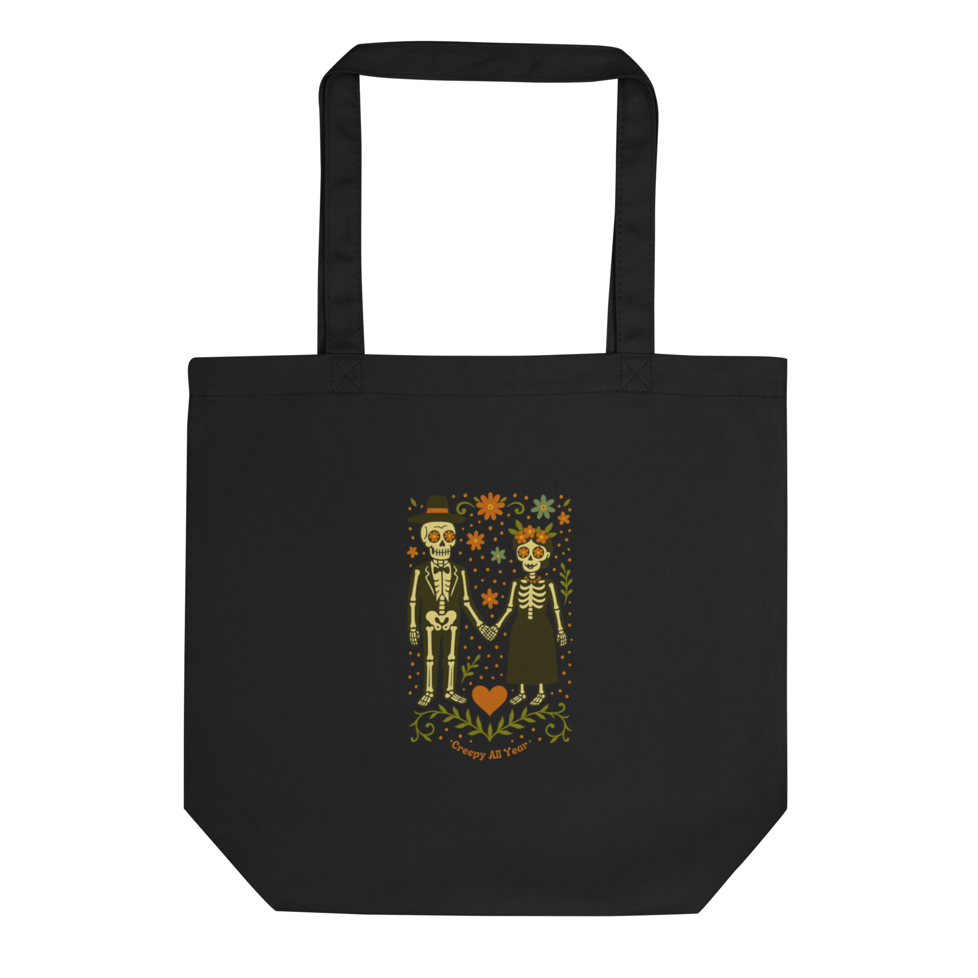 This is a black tote bag with a Día de los Muertos–style illustration of a male and female skeleton holding hands, surrounded by marigolds, vines, and folk art patterns. The skeletons wear traditional black clothing, the man in a suit and hat, the woman with a floral crown with a red heart and leaves at the bottom and the text Creepy All Year. Rendered in warm tones of orange, green, and cream, symbolizing eternal love and remembrance