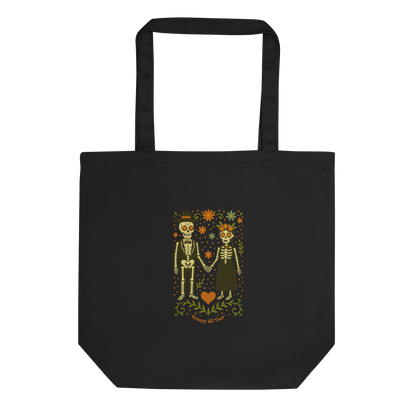 This is a black tote bag with a Día de los Muertos–style illustration of a male and female skeleton holding hands, surrounded by marigolds, vines, and folk art patterns. The skeletons wear traditional black clothing, the man in a suit and hat, the woman with a floral crown with a red heart and leaves at the bottom and the text Creepy All Year. Rendered in warm tones of orange, green, and cream, symbolizing eternal love and remembrance