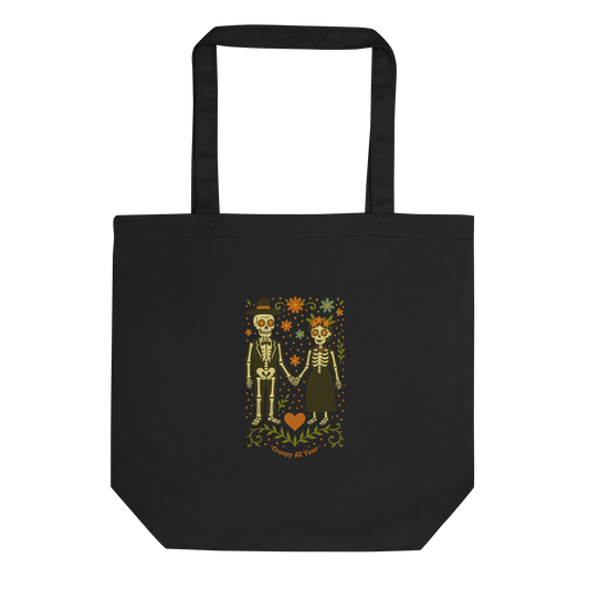 This is a black tote bag with a Día de los Muertos–style illustration of a male and female skeleton holding hands, surrounded by marigolds, vines, and folk art patterns. The skeletons wear traditional black clothing, the man in a suit and hat, the woman with a floral crown with a red heart and leaves at the bottom and the text Creepy All Year. Rendered in warm tones of orange, green, and cream, symbolizing eternal love and remembrance