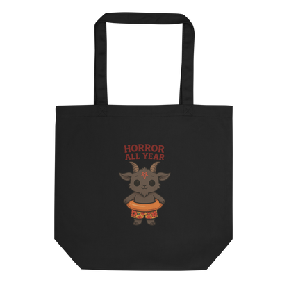 This is a black tote bag that has a cartoon illustration of a baby Baphomet goat with curved horns and a playful grin, wearing bright floral swim trunks while floating in a colorful inner tube. The character is set against a light background with the bold text “Horror All Year” above in red letters 