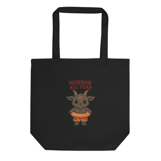 This is a black tote bag that has a cartoon illustration of a baby Baphomet goat with curved horns and a playful grin, wearing bright floral swim trunks while floating in a colorful inner tube. The character is set against a light background with the bold text “Horror All Year” above in red letters 