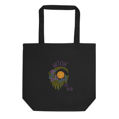 This is a black eco tote bag with a design of a hand-drawn wreath made of moss-covered branches wraps around a glowing sun. The wreath is decorated with purple and blue flowers, small amethyst and fluorite crystals, and strands of hanging moss. The words “Witchy” curve above the wreath and “All Year” sits on the bottom right 