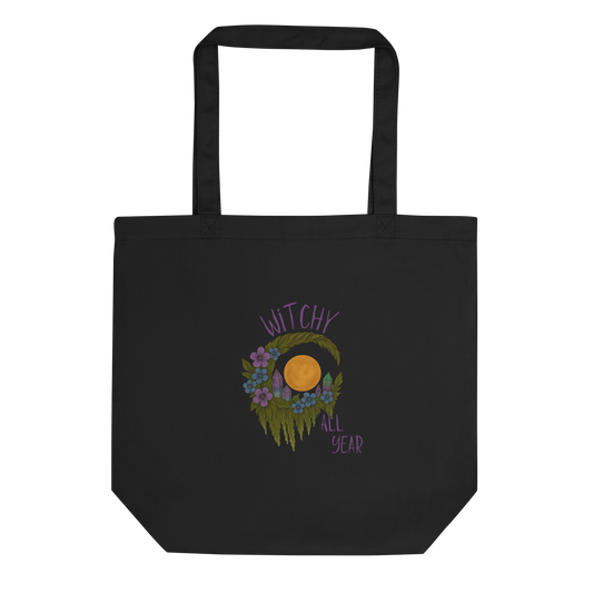This is a black eco tote bag with a design of a hand-drawn wreath made of moss-covered branches wraps around a glowing sun. The wreath is decorated with purple and blue flowers, small amethyst and fluorite crystals, and strands of hanging moss. The words “Witchy” curve above the wreath and “All Year” sits on the bottom right 