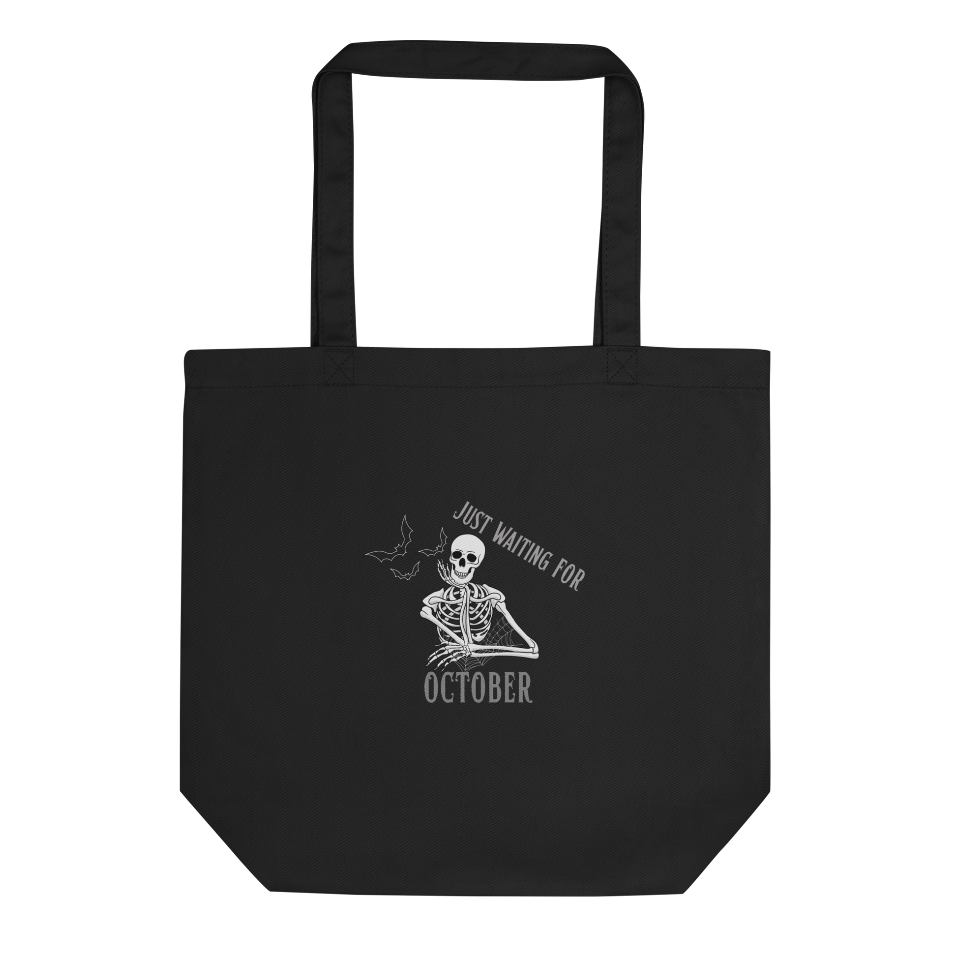This is a black tote bag that has a bored skeleton with his head resting in his hand and he has spider webs and bats around him and the text reads just waiting for October