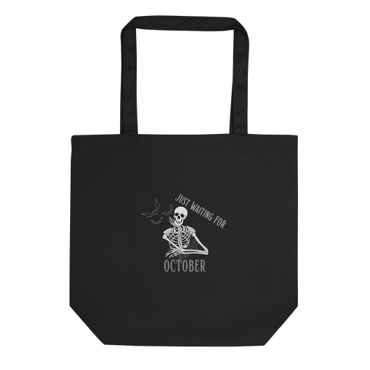 This is a black tote bag that has a bored skeleton with his head resting in his hand and he has spider webs and bats around him and the text reads just waiting for October
