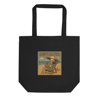 This is a eco tote bag with a retro-style illustration of a grinning skeleton lounging on a beach chair under an orange umbrella. The skeleton is holding a pumpkin-shaped drink cup with a straw and relaxing by the ocean, with a bright sun in the sky and a life preserver in the sand. The words “Creepy All Year” appear boldly at the top 