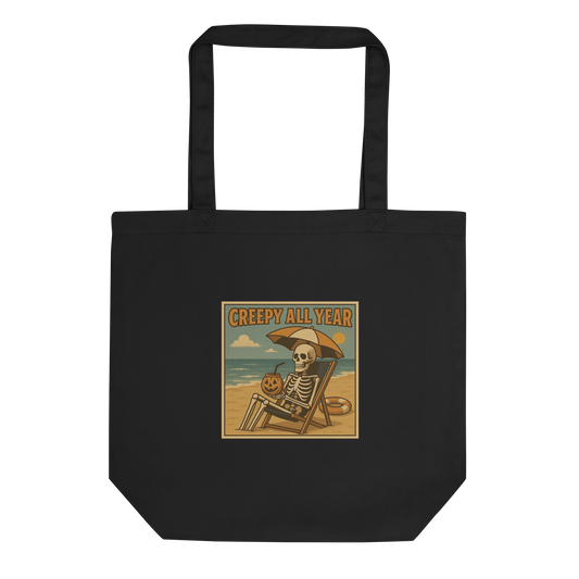 This is a eco tote bag with a retro-style illustration of a grinning skeleton lounging on a beach chair under an orange umbrella. The skeleton is holding a pumpkin-shaped drink cup with a straw and relaxing by the ocean, with a bright sun in the sky and a life preserver in the sand. The words “Creepy All Year” appear boldly at the top 