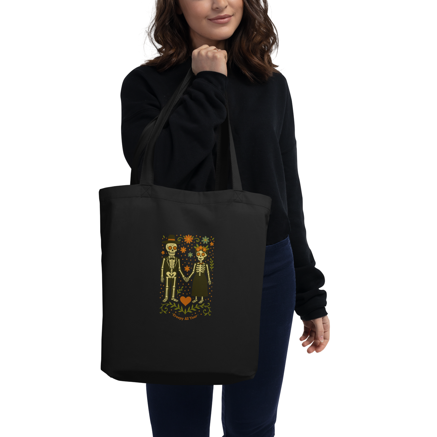 This is a woman with dark hair holding a black tote bag with a Día de los Muertos–style illustration of a male and female skeleton holding hands, surrounded by marigolds, vines, and folk art patterns. The skeletons wear traditional black clothing, the man in a suit and hat, the woman with a floral crown with a red heart and leaves at the bottom and the text Creepy All Year. Rendered in warm tones of orange, green, and cream, symbolizing eternal love and remembrance
