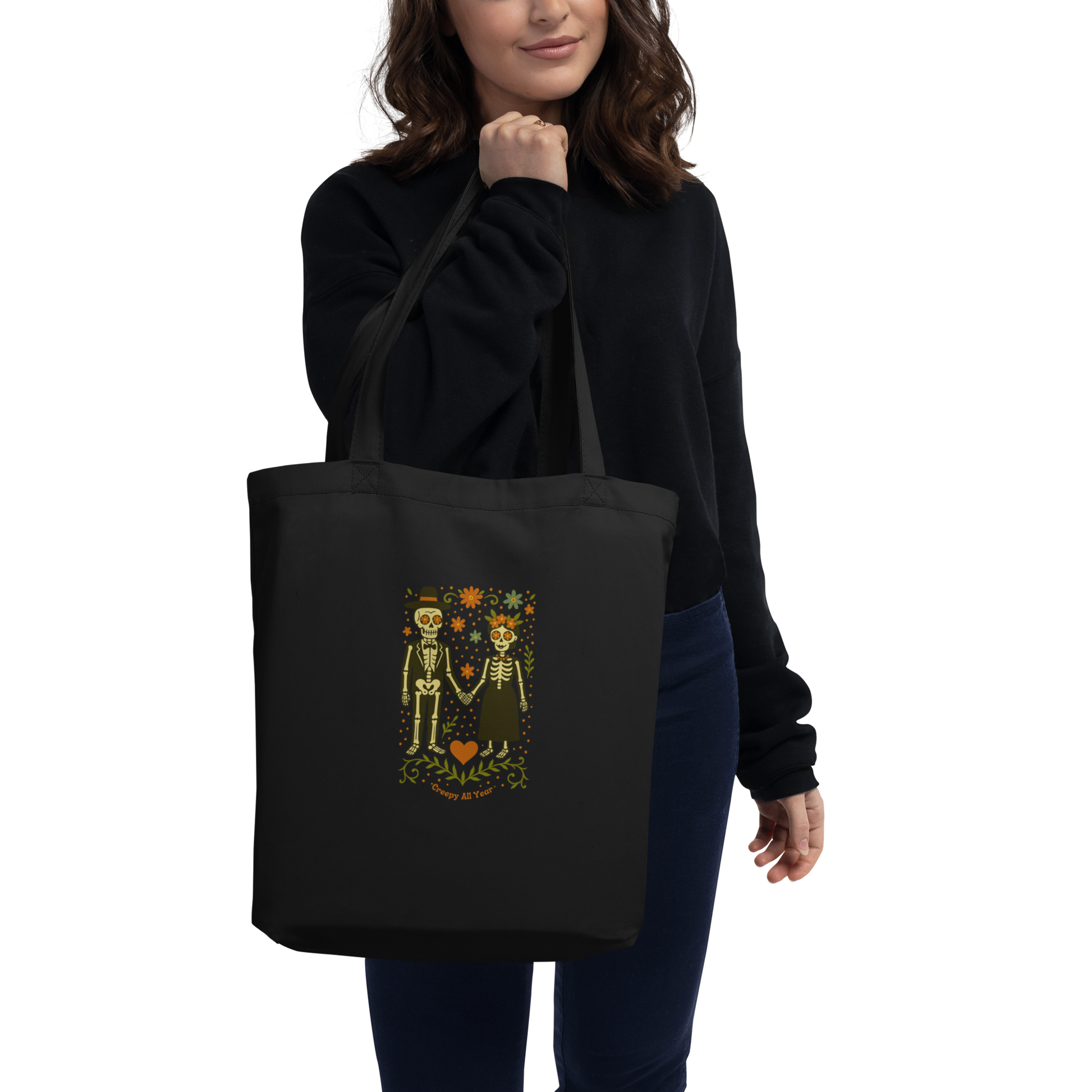 This is a woman with dark hair holding a black tote bag with a Día de los Muertos–style illustration of a male and female skeleton holding hands, surrounded by marigolds, vines, and folk art patterns. The skeletons wear traditional black clothing, the man in a suit and hat, the woman with a floral crown with a red heart and leaves at the bottom and the text Creepy All Year. Rendered in warm tones of orange, green, and cream, symbolizing eternal love and remembrance