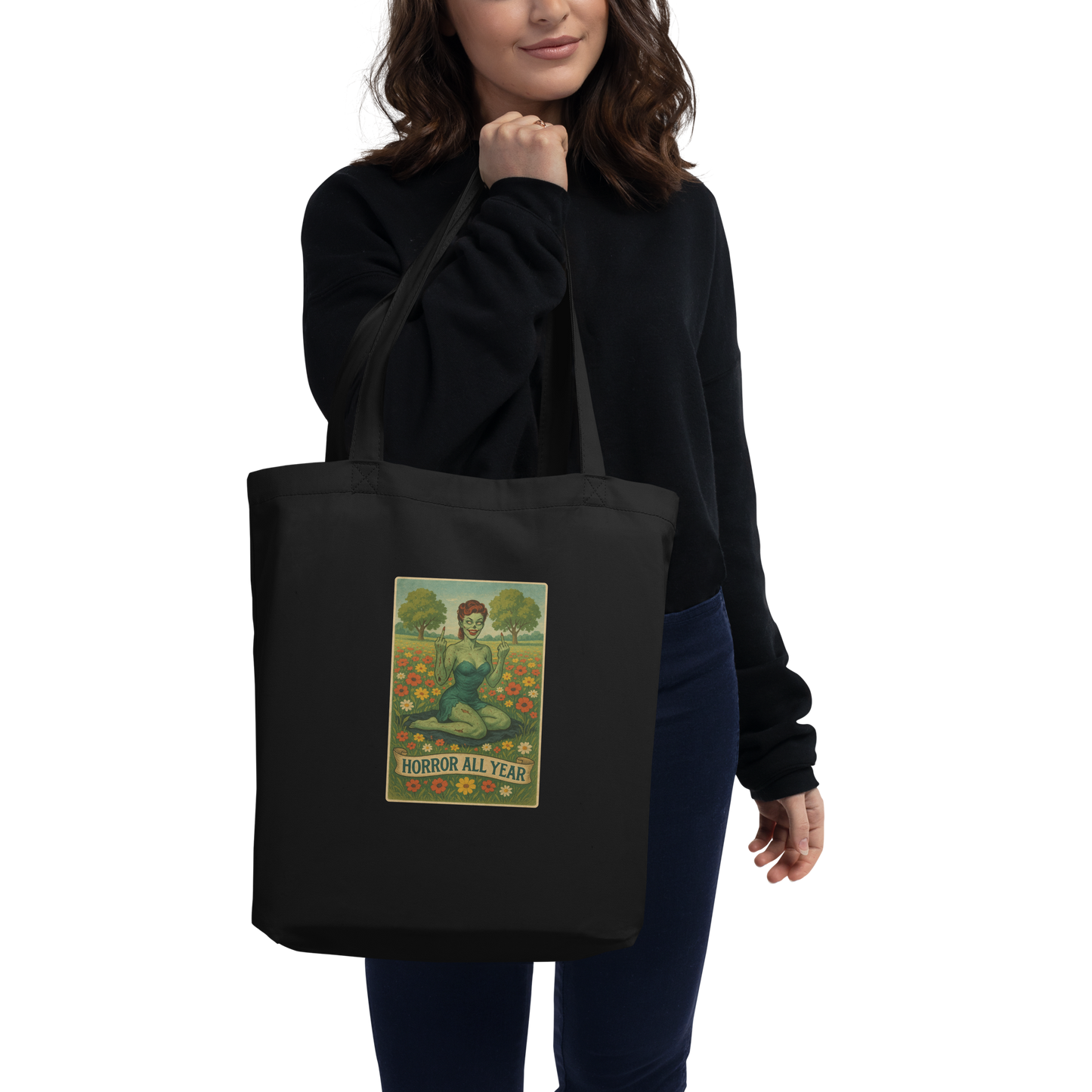 This is a woman with dark hair holding a black eco tote bag with a design illustration of a sassy zombie pinup girl standing in a floral field, wearing tattered lingerie and flipping off the viewer with both hands. She has green skin, vintage curls, and a bold attitude. The design blends horror and humor, with a pastel color palette and creepy cute vibes