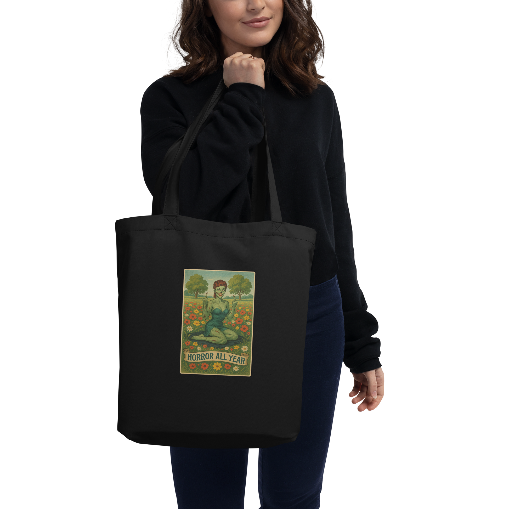 This is a woman with dark hair holding a black eco tote bag with a design illustration of a sassy zombie pinup girl standing in a floral field, wearing tattered lingerie and flipping off the viewer with both hands. She has green skin, vintage curls, and a bold attitude. The design blends horror and humor, with a pastel color palette and creepy cute vibes