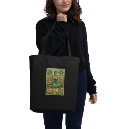 This is a woman with dark hair holding a black eco tote bag with a design illustration of a sassy zombie pinup girl standing in a floral field, wearing tattered lingerie and flipping off the viewer with both hands. She has green skin, vintage curls, and a bold attitude. The design blends horror and humor, with a pastel color palette and creepy cute vibes