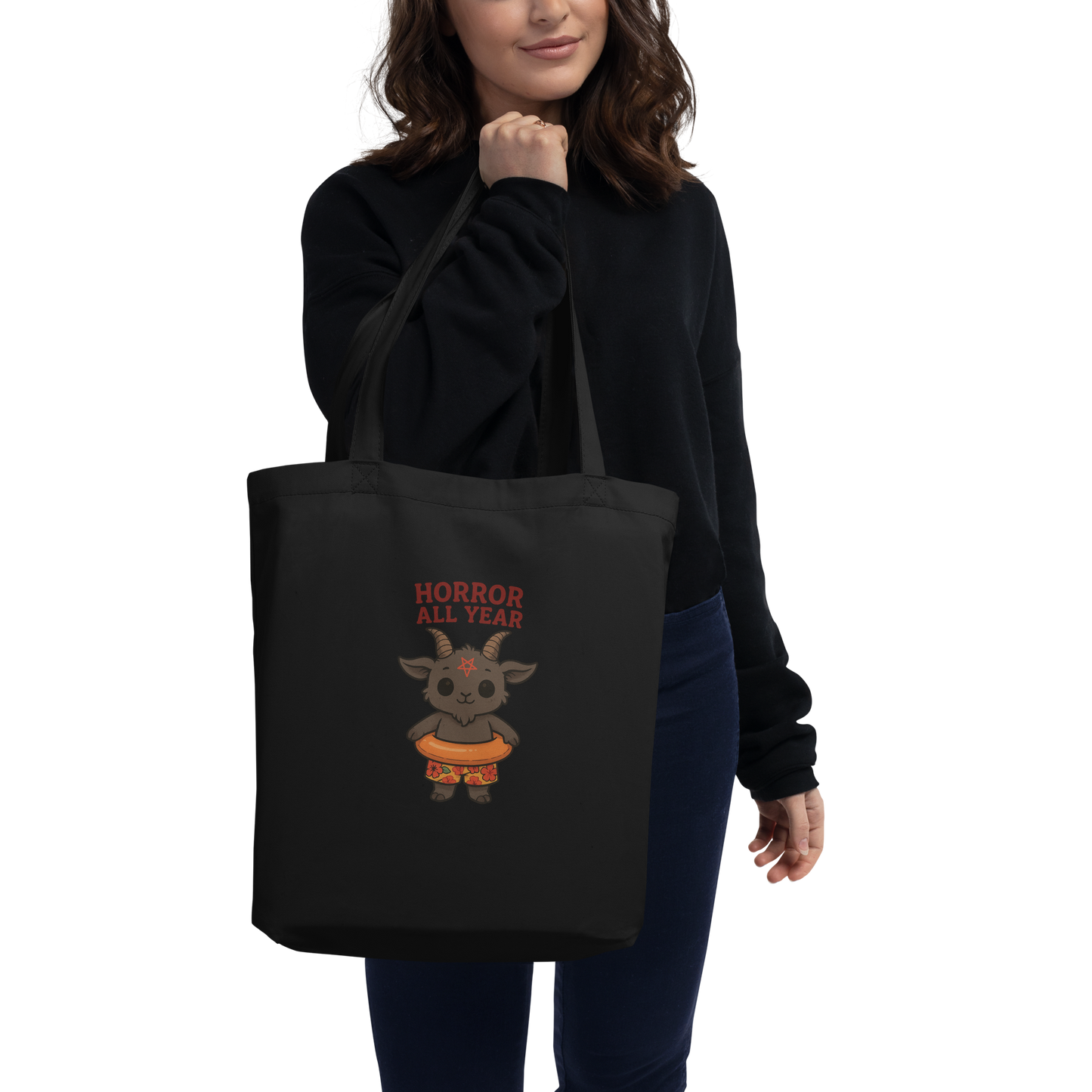 This is a woman holding a black tote bag that has a cartoon illustration of a baby Baphomet goat with curved horns and a playful grin, wearing bright floral swim trunks while floating in a colorful inner tube. The character is set against a light background with the bold text “Horror All Year” above in red letters 
