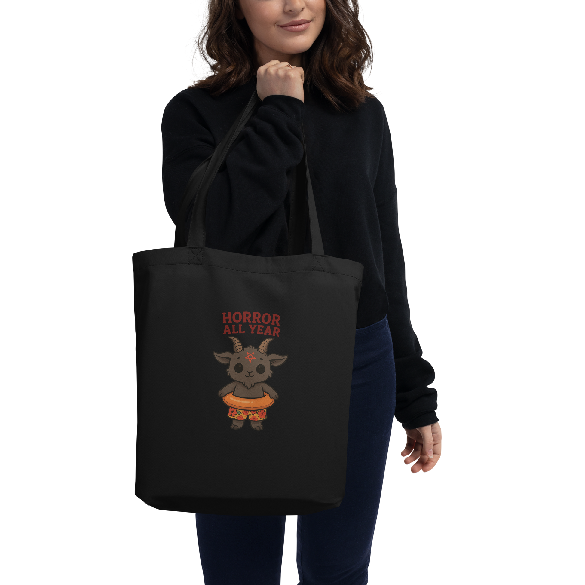 This is a woman holding a black tote bag that has a cartoon illustration of a baby Baphomet goat with curved horns and a playful grin, wearing bright floral swim trunks while floating in a colorful inner tube. The character is set against a light background with the bold text “Horror All Year” above in red letters 