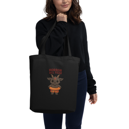 This is a woman holding a black tote bag that has a cartoon illustration of a baby Baphomet goat with curved horns and a playful grin, wearing bright floral swim trunks while floating in a colorful inner tube. The character is set against a light background with the bold text “Horror All Year” above in red letters 