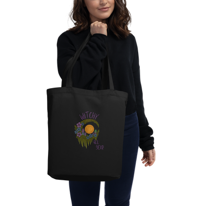 This is a woman with dark hair holding a black eco tote bag with a design of a hand-drawn wreath made of moss-covered branches wraps around a glowing sun. The wreath is decorated with purple and blue flowers, small amethyst and fluorite crystals, and strands of hanging moss. The words “Witchy” curve above the wreath and “All Year” sits on the bottom right 