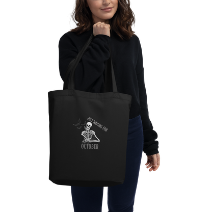 This is a woman with dark hair holding a black tote bag that has a bored skeleton with his head resting in his hand and he has spider webs and bats around him and the text reads just waiting for October