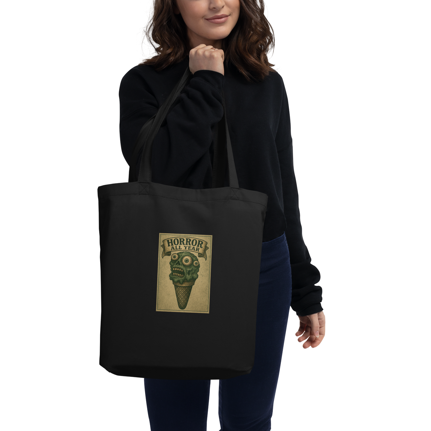 This is a woman with dark hair holding a black tote bag  that is a vintage-style illustration of a zombie-themed ice cream cone with a green, melting undead face and bulging eyeballs. Above the cone, a horror banner reads "Horror All Year" on a distressed parchment background