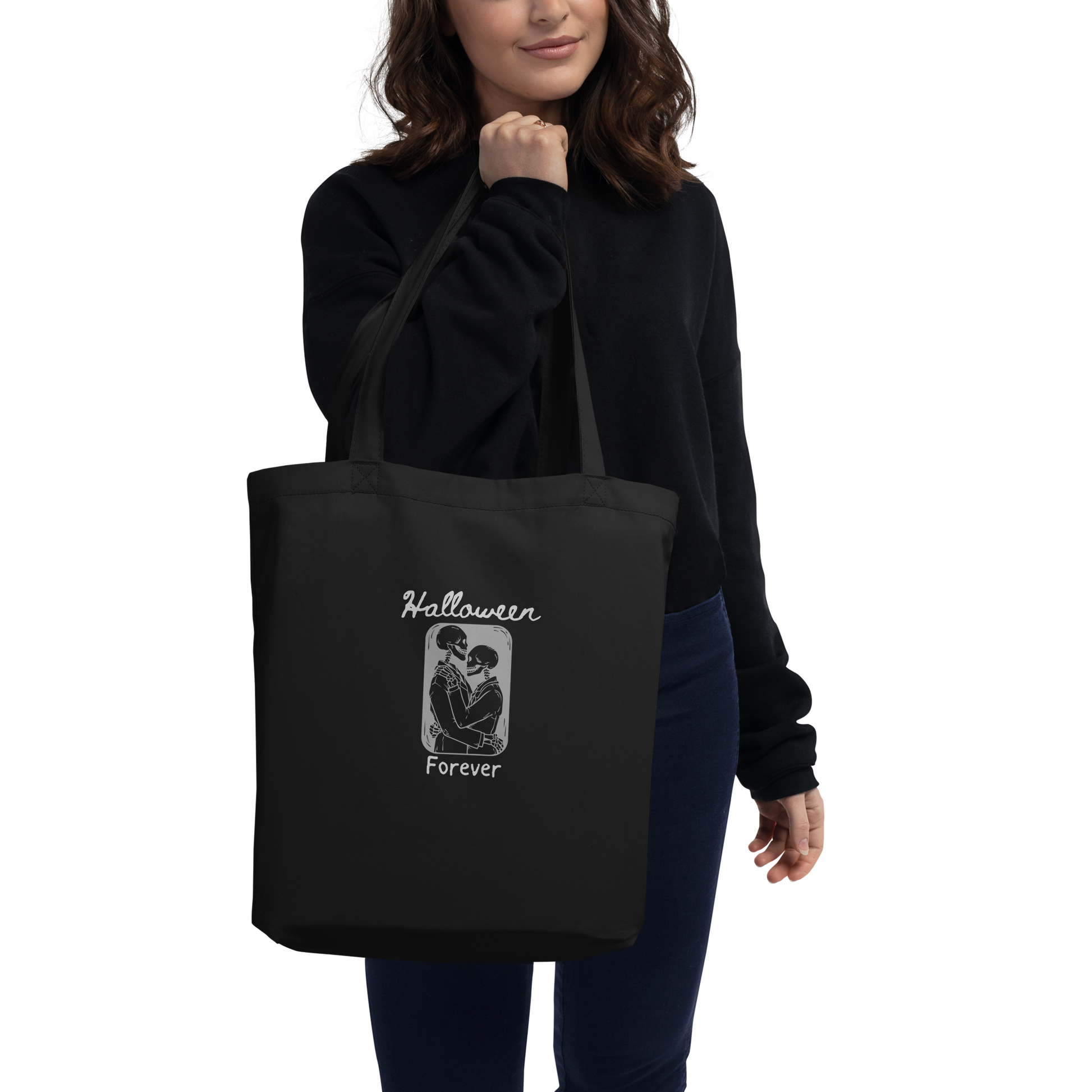 This is a woman holding a black tote bag that has a hugging skeleton couple that is grey and says Halloween Forever 
