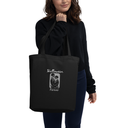 This is a woman holding a black tote bag that has a hugging skeleton couple that is grey and says Halloween Forever 