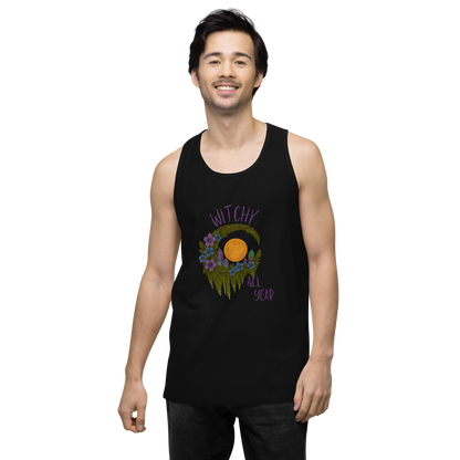This is a man with dark hair wearing a black tank top with a design of a hand-drawn wreath made of moss-covered branches wraps around a glowing sun. The wreath is decorated with purple and blue flowers, small amethyst and fluorite crystals, and strands of hanging moss. The words “Witchy” curve above the wreath and “All Year” sits on the bottom right 
