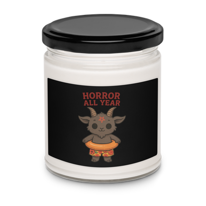 This is a white candle with a black tin lid on top and on the front is a cartoon illustration of a baby Baphomet goat with curved horns and a playful grin, wearing bright floral swim trunks while floating in a colorful inner tube. The character is set against a light background with the bold text “Horror All Year” above in red letters