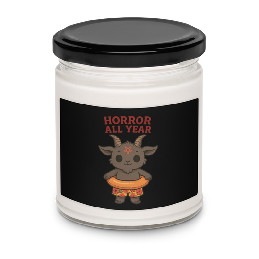 This is a white candle with a black tin lid on top and on the front is a cartoon illustration of a baby Baphomet goat with curved horns and a playful grin, wearing bright floral swim trunks while floating in a colorful inner tube. The character is set against a light background with the bold text “Horror All Year” above in red letters