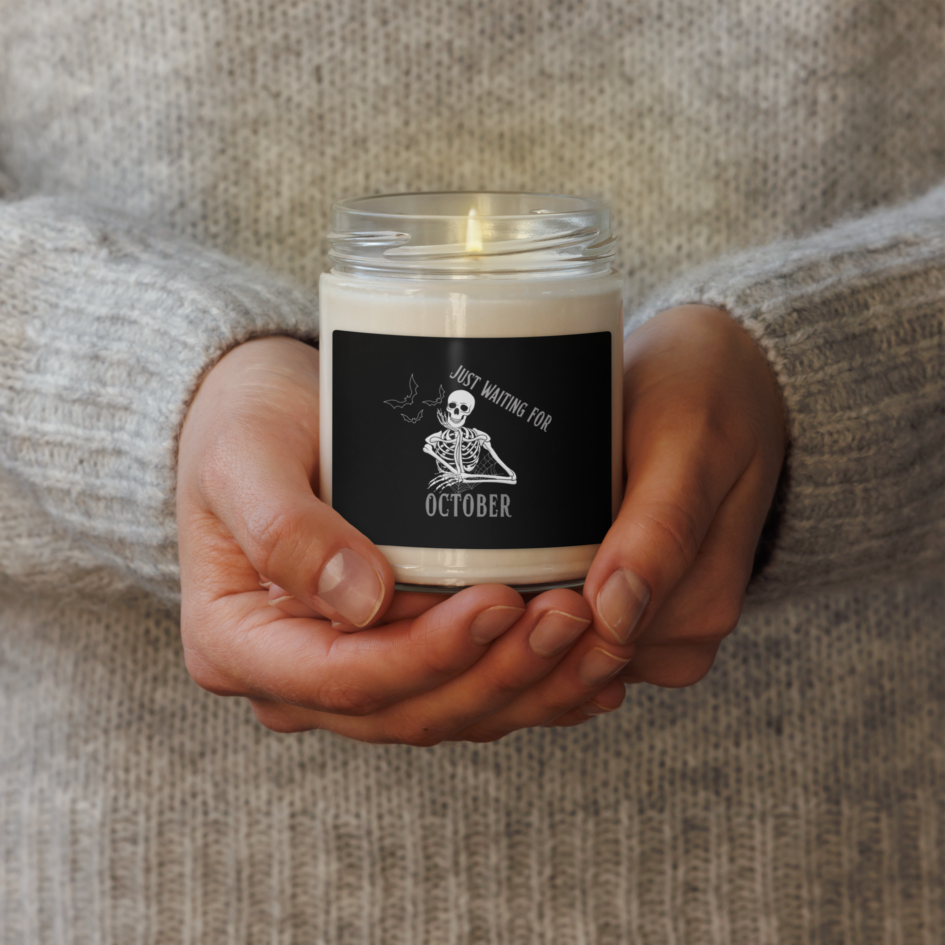 This is woman holding a white candle with black label that has a bored skeleton with his head resting in his hand and he has spider webs and bats around him and the text reads just waiting for October
