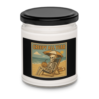This is a white candle with black tin lid and black label with a retro-style illustration of a grinning skeleton lounging on a beach chair under an orange umbrella. The skeleton is holding a pumpkin-shaped drink cup with a straw and relaxing by the ocean, with a bright sun in the sky and a life preserver in the sand. The words “Creepy All Year” appear boldly at the top 