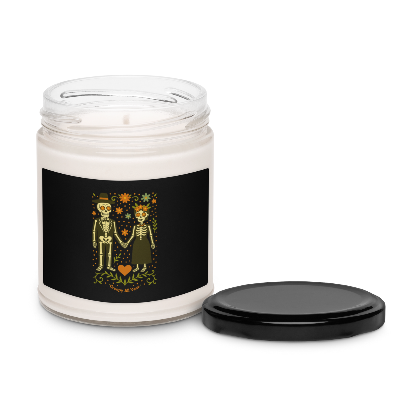 This is a white candle and black tin lid with a black with a Día de los Muertos–style illustration of a male and female skeleton holding hands, surrounded by marigolds, vines, and folk art patterns. The skeletons wear traditional black clothing, the man in a suit and hat, the woman with a floral crown with a red heart and leaves at the bottom and the text Creepy All Year. Rendered in warm tones of orange, green, and cream, symbolizing eternal love and remembrance