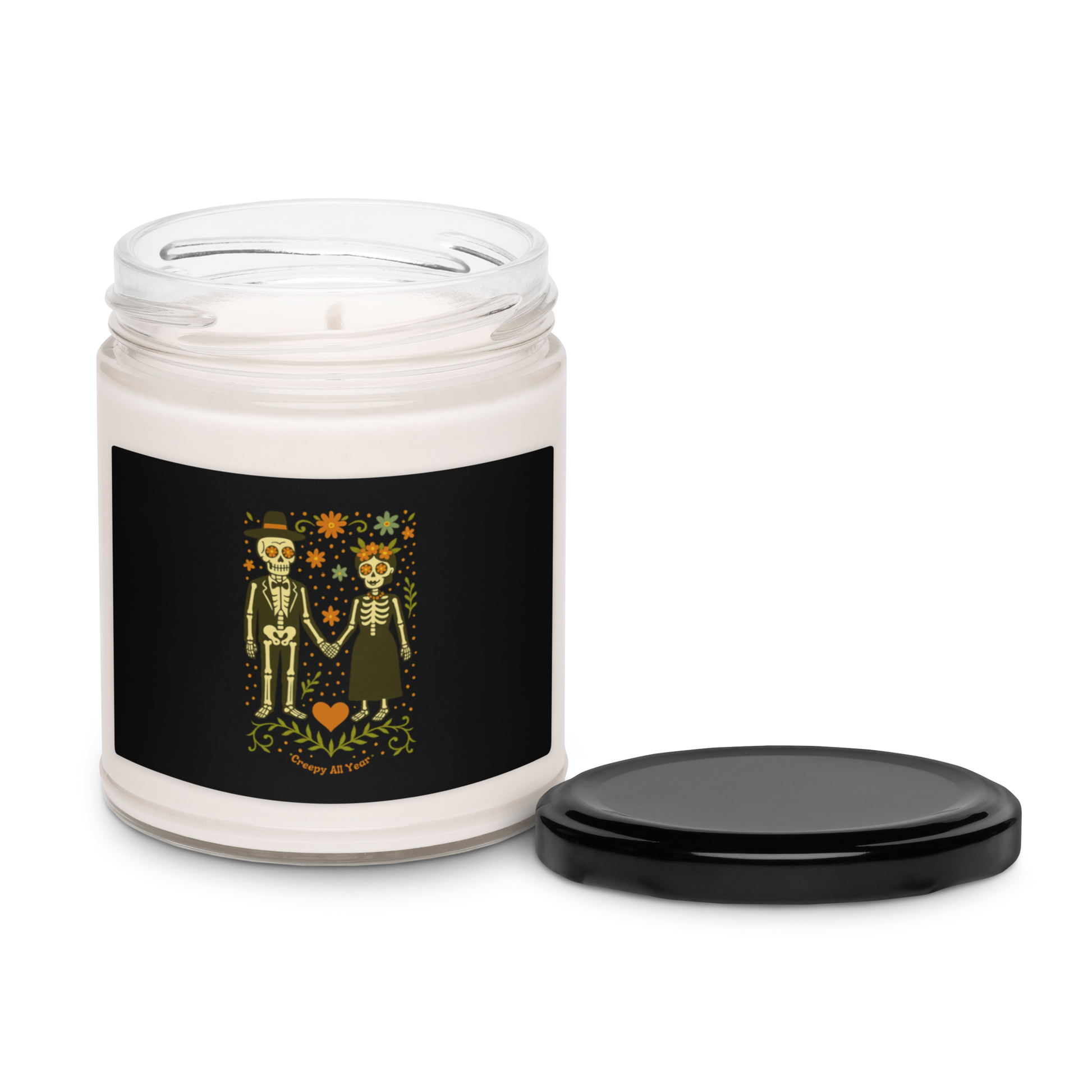 This is a white candle and black tin lid with a black with a Día de los Muertos–style illustration of a male and female skeleton holding hands, surrounded by marigolds, vines, and folk art patterns. The skeletons wear traditional black clothing, the man in a suit and hat, the woman with a floral crown with a red heart and leaves at the bottom and the text Creepy All Year. Rendered in warm tones of orange, green, and cream, symbolizing eternal love and remembrance