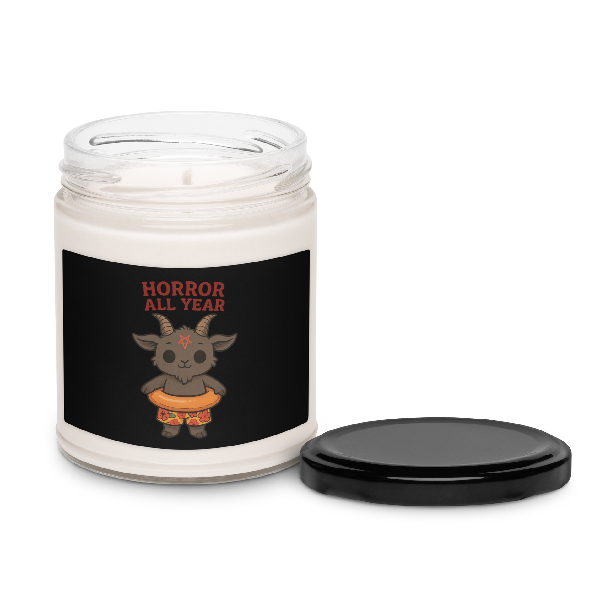 This is a white candle with a black tin lid on the front has a cartoon illustration of a baby Baphomet goat with curved horns and a playful grin, wearing bright floral swim trunks while floating in a colorful inner tube. The character is set against a light background with the bold text “Horror All Year” above in red letters