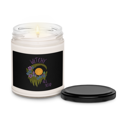 This is a white candle and black tin lid with a design of a hand-drawn wreath made of moss-covered branches wraps around a glowing sun. The wreath is decorated with purple and blue flowers, small amethyst and fluorite crystals, and strands of hanging moss. The words “Witchy” curve above the wreath and “All Year” sits on the bottom right 