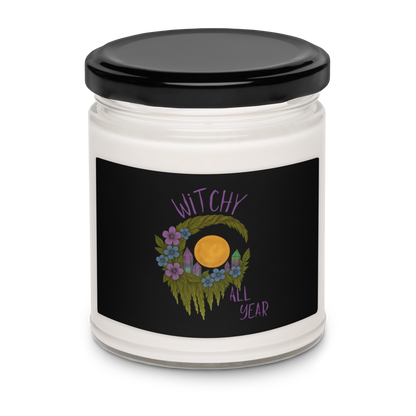 This is a white candle with a design of a hand-drawn wreath made of moss-covered branches wraps around a glowing sun. The wreath is decorated with purple and blue flowers, small amethyst and fluorite crystals, and strands of hanging moss. The words “Witchy” curve above the wreath and “All Year” sits on the bottom right 