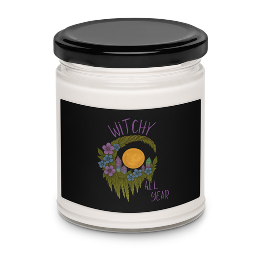 This is a white candle with a design of a hand-drawn wreath made of moss-covered branches wraps around a glowing sun. The wreath is decorated with purple and blue flowers, small amethyst and fluorite crystals, and strands of hanging moss. The words “Witchy” curve above the wreath and “All Year” sits on the bottom right 