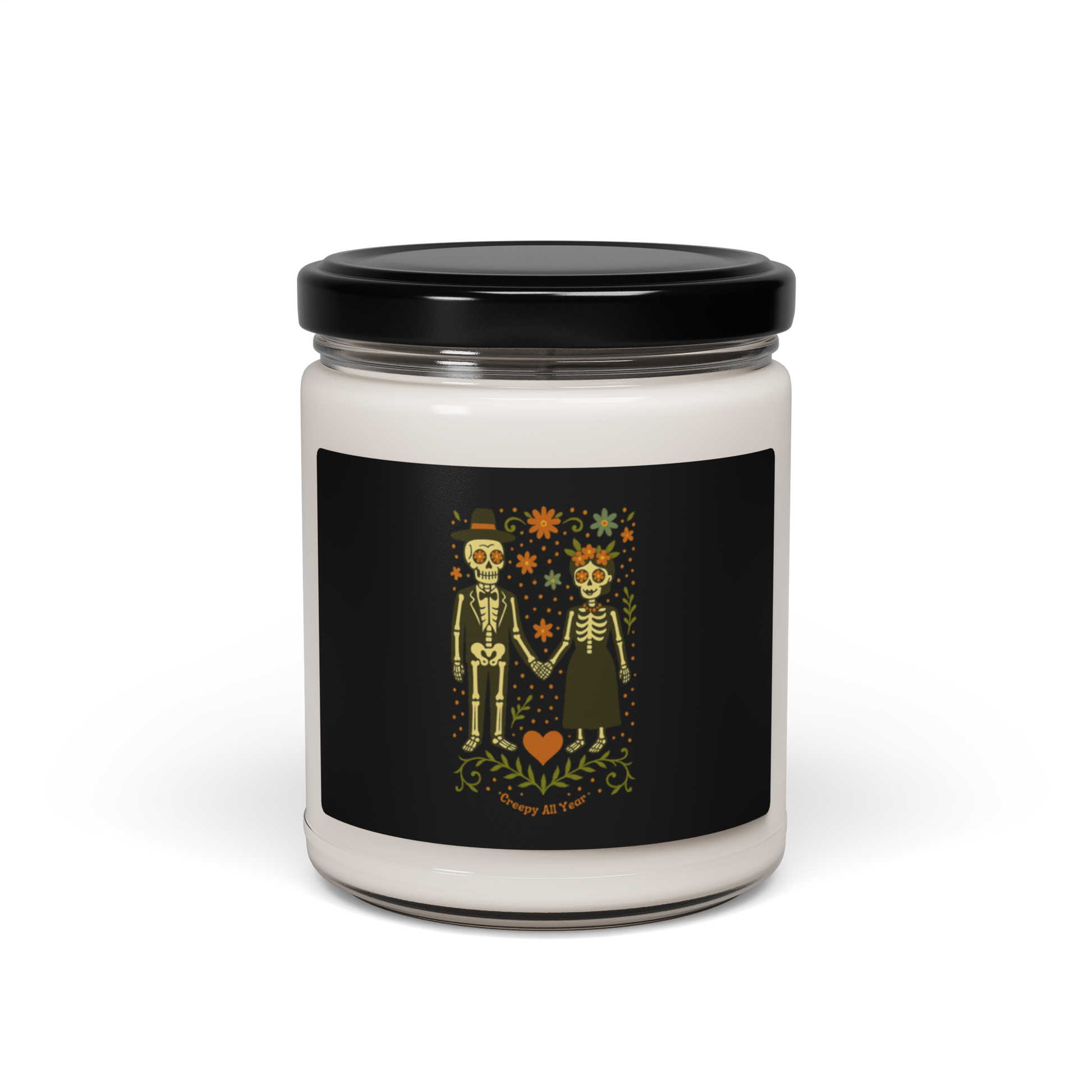 This is a white candle with a black with a Día de los Muertos–style illustration of a male and female skeleton holding hands, surrounded by marigolds, vines, and folk art patterns. The skeletons wear traditional black clothing, the man in a suit and hat, the woman with a floral crown with a red heart and leaves at the bottom and the text Creepy All Year. Rendered in warm tones of orange, green, and cream, symbolizing eternal love and remembrance