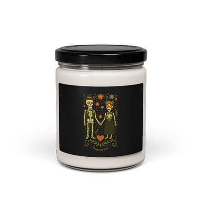 This is a white candle with a black with a Día de los Muertos–style illustration of a male and female skeleton holding hands, surrounded by marigolds, vines, and folk art patterns. The skeletons wear traditional black clothing, the man in a suit and hat, the woman with a floral crown with a red heart and leaves at the bottom and the text Creepy All Year. Rendered in warm tones of orange, green, and cream, symbolizing eternal love and remembrance