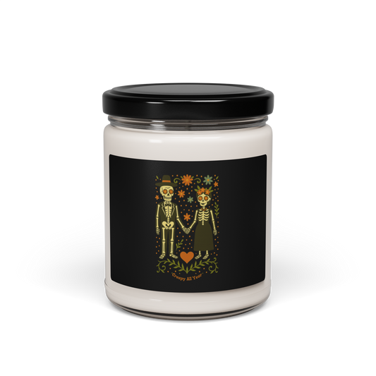 This is a white candle with a black with a Día de los Muertos–style illustration of a male and female skeleton holding hands, surrounded by marigolds, vines, and folk art patterns. The skeletons wear traditional black clothing, the man in a suit and hat, the woman with a floral crown with a red heart and leaves at the bottom and the text Creepy All Year. Rendered in warm tones of orange, green, and cream, symbolizing eternal love and remembrance