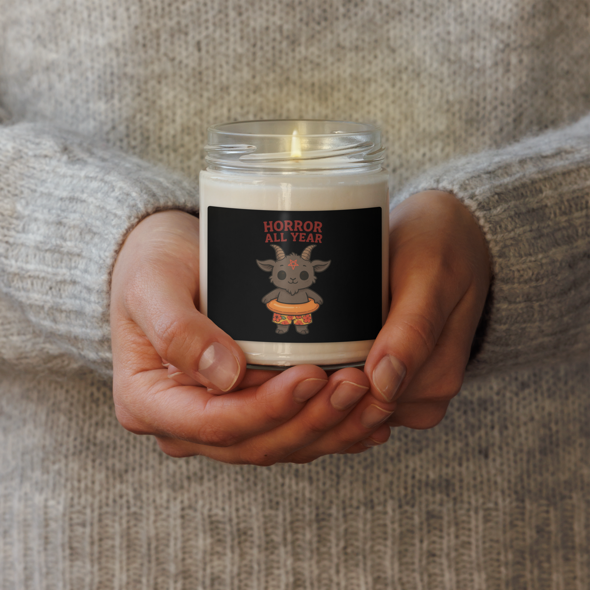 This is a woman holding a white candle with a cartoon illustration of a baby Baphomet goat with curved horns and a playful grin, wearing bright floral swim trunks while floating in a colorful inner tube. The character is set against a light background with the bold text “Horror All Year” above in red letters