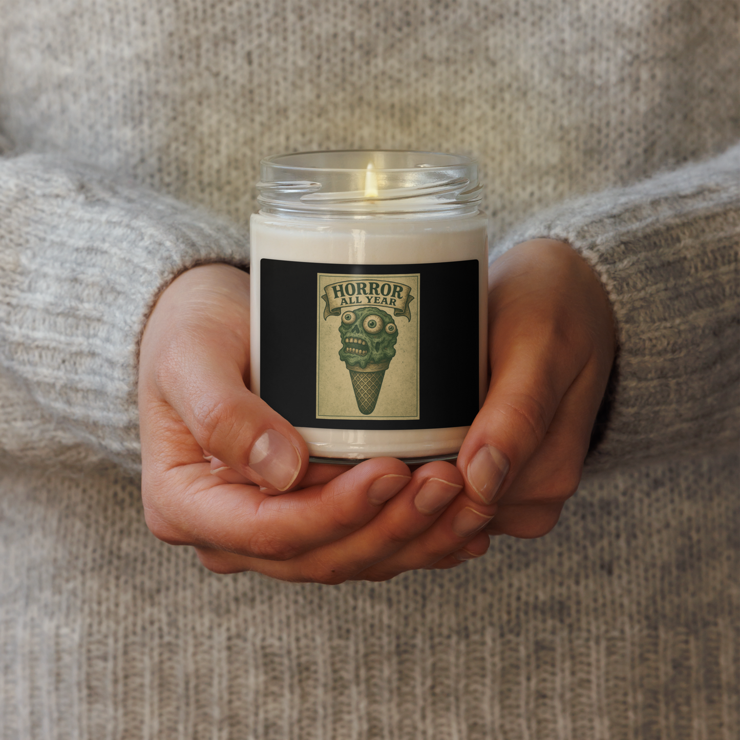This is a woman holding a candle with a black label that is a vintage-style illustration of a zombie-themed ice cream cone with a green, melting undead face and bulging eyeballs. Above the cone, a horror banner reads "Horror All Year" on a distressed parchment background