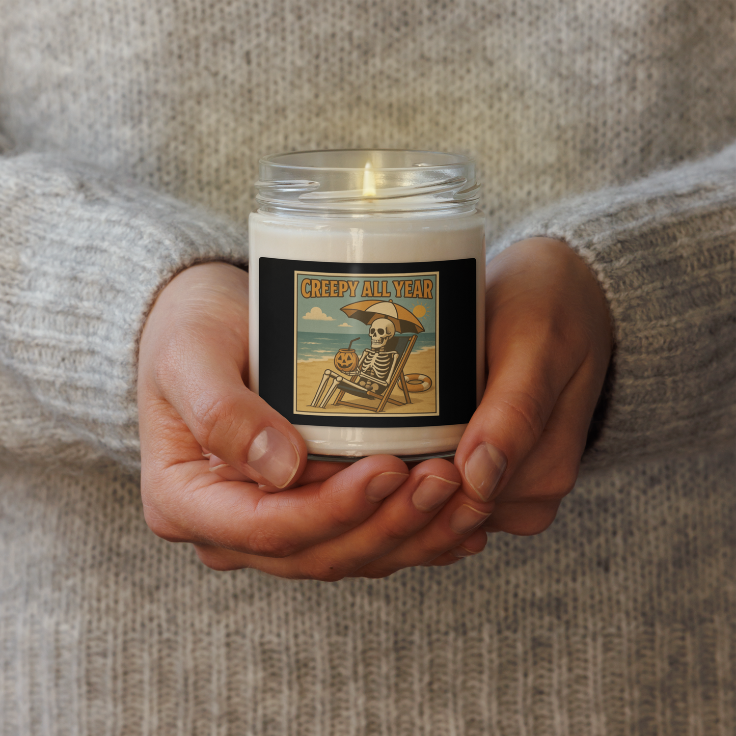 This is a woman's hands holding a white candle with black tin lid and black label with a retro-style illustration of a grinning skeleton lounging on a beach chair under an orange umbrella. The skeleton is holding a pumpkin-shaped drink cup with a straw and relaxing by the ocean, with a bright sun in the sky and a life preserver in the sand. The words “Creepy All Year” appear boldly at the top 