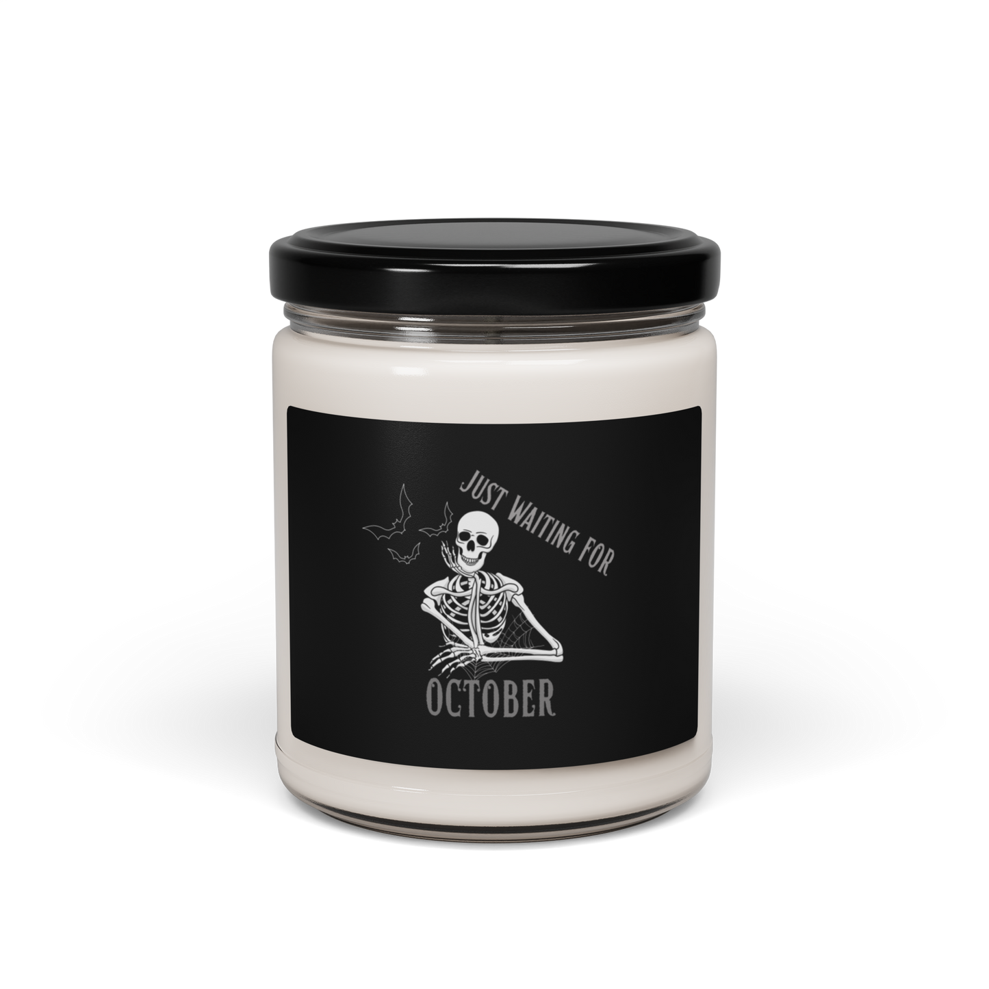 This is a white candle with black label that has a bored skeleton with his head resting in his hand and he has spider webs and bats around him and the text reads just waiting for October