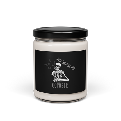 This is a white candle with black label that has a bored skeleton with his head resting in his hand and he has spider webs and bats around him and the text reads just waiting for October