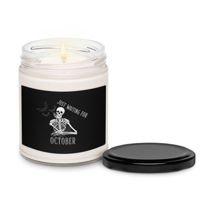 This is a white candle with black lid and label that has a bored skeleton with his head resting in his hand and he has spider webs and bats around him and the text reads just waiting for October