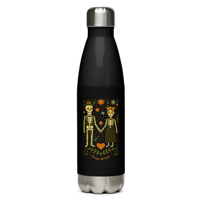 This is a black and stainless steel water bottle with a Día de los Muertos–style illustration of a male and female skeleton holding hands, surrounded by marigolds, vines, and folk art patterns. The skeletons wear traditional black clothing, the man in a suit and hat, the woman with a floral crown with a red heart and leaves at the bottom and the text Creepy All Year. Rendered in warm tones of orange, green, and cream, symbolizing eternal love and remembrance