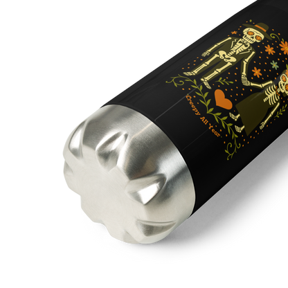 This is a zoom in of a black and stainless steel water bottle with a Día de los Muertos–style illustration of a male and female skeleton holding hands, surrounded by marigolds, vines, and folk art patterns. The skeletons wear traditional black clothing, the man in a suit and hat, the woman with a floral crown with a red heart and leaves at the bottom and the text Creepy All Year. Rendered in warm tones of orange, green, and cream, symbolizing eternal love and remembrance