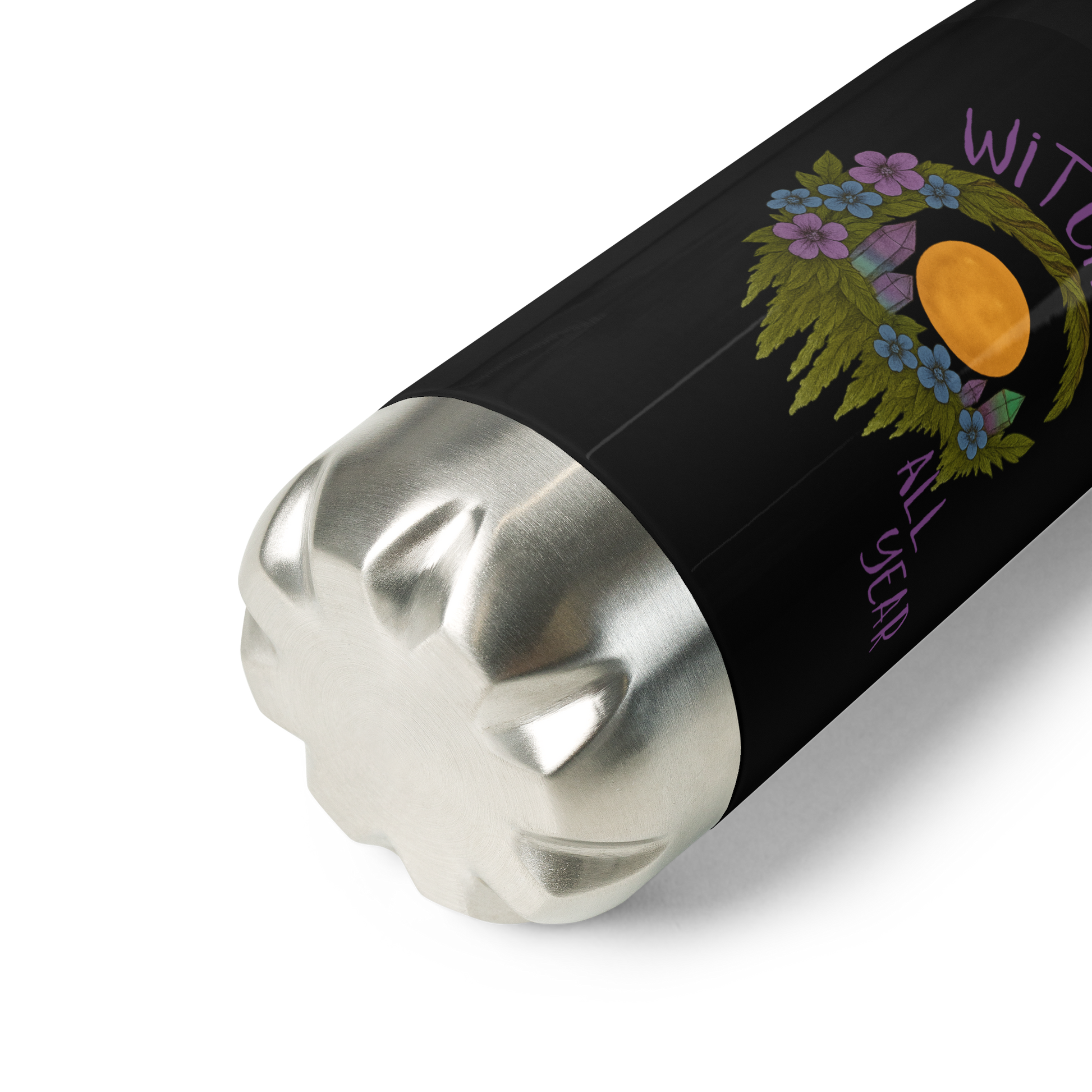 This is a zoom in of a black stainless steel water bottle  with a design of a hand-drawn wreath made of moss-covered branches wraps around a glowing sun. The wreath is decorated with purple and blue flowers, small amethyst and fluorite crystals, and strands of hanging moss. The words “Witchy” curve above the wreath and “All Year” sits on the bottom right 